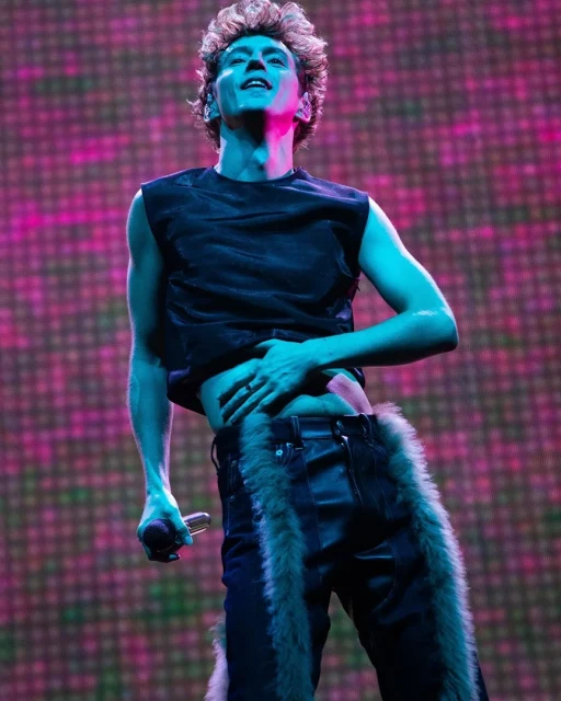 Musician performing on stage with colorful background lights, wearing black top and black pants with fur trim and holding a microphone.