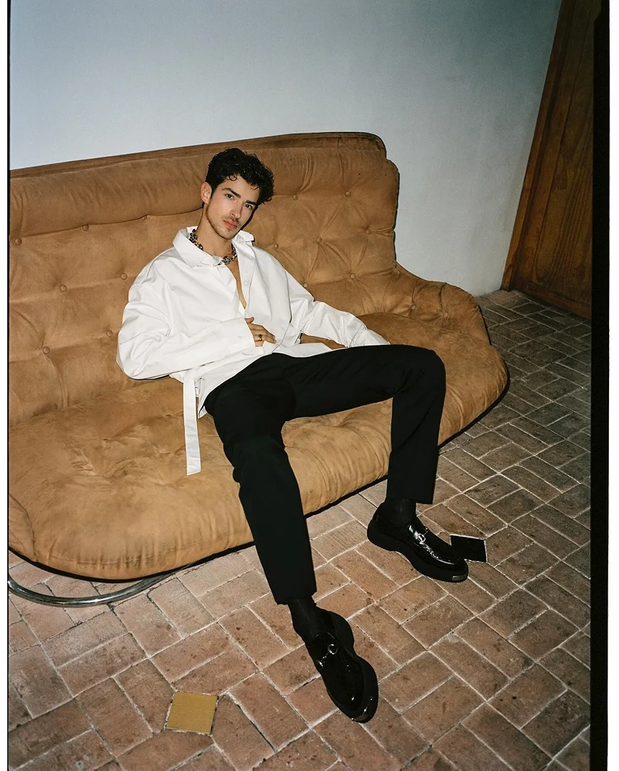 A young adult male sits casually on a vintage couch in a fashion style, wearing a white shirt, black pants, and shiny shoes. 