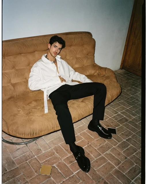 A young adult male sits casually on a vintage couch in a fashion style, wearing a white shirt, black pants, and shiny shoes. 