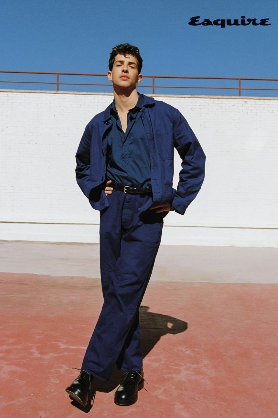 A young man in a blue work suit poses against a white wall and blue sky. He wears black dress shoes and has his hands on his hips.