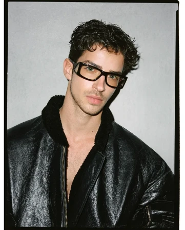 Close-up portrait of a man in a black leather jacket and thick black frame eyeglasses against a neutral gray background.