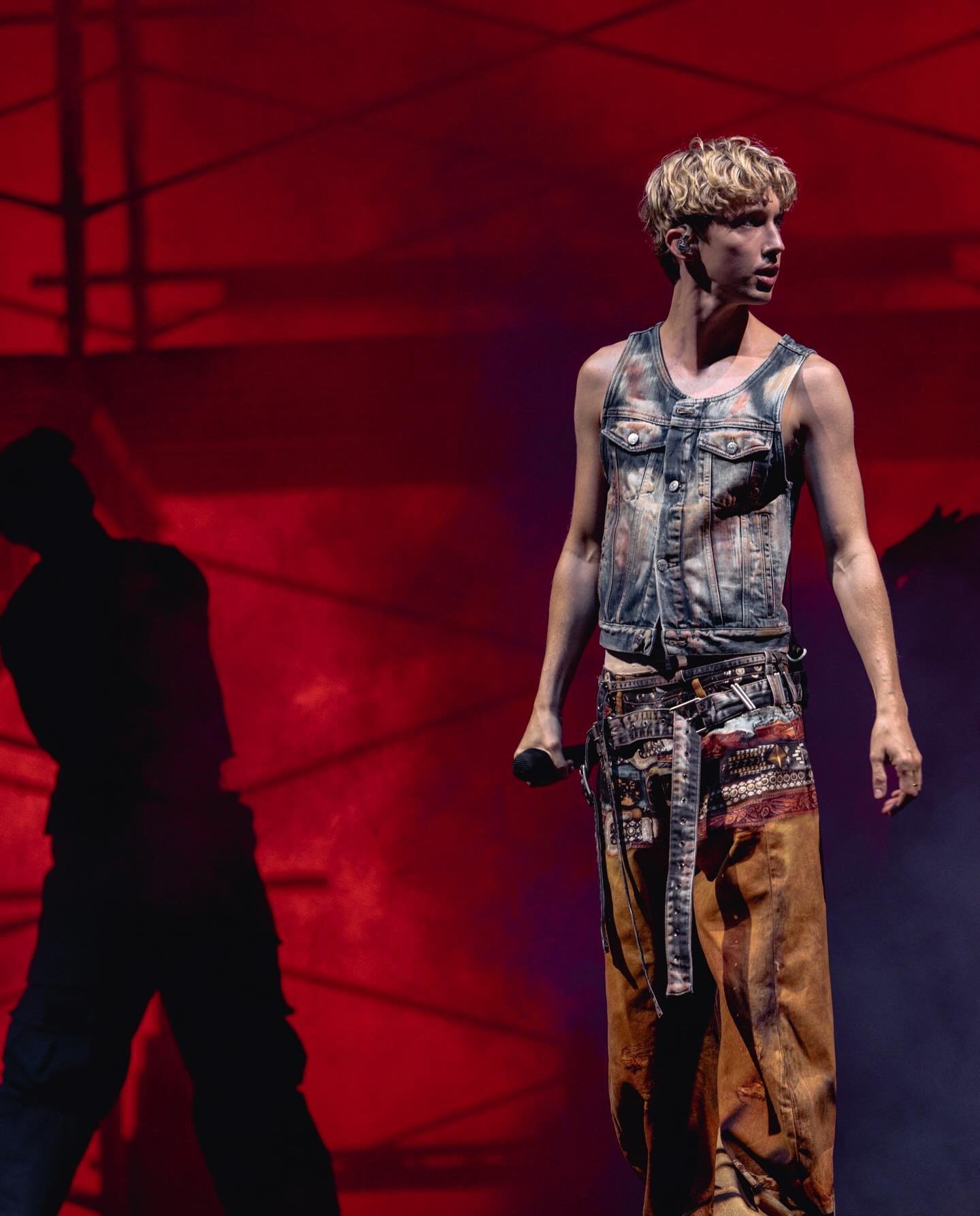Male singer performs on a red-lit stage wearing denim vest, patterned belt and brown pants with a shadow cast on the backdrop.