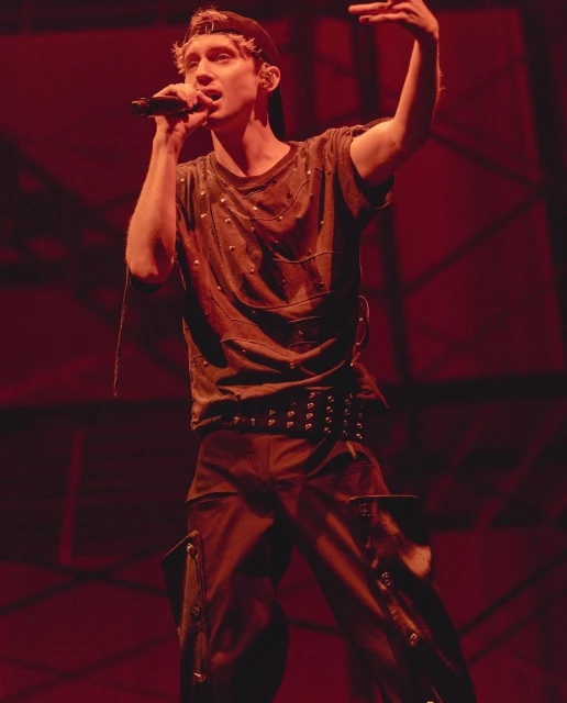 Singer performing on stage, holding microphone, wearing t-shirt, trousers with studded belt and baseball cap in red stage lighting.