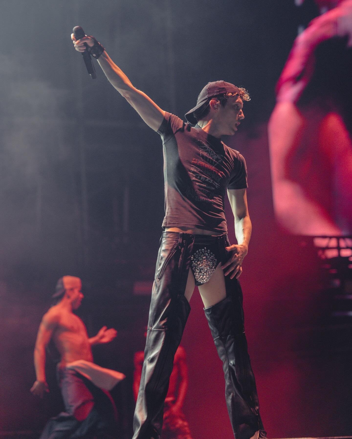 Live music performance featuring a male singer in a cap, t-shirt, and chaps, holding a microphone on stage, illuminated by red lighting.
