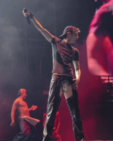 Live music performance featuring a male singer in a cap, t-shirt, and chaps, holding a microphone on stage, illuminated by red lighting.