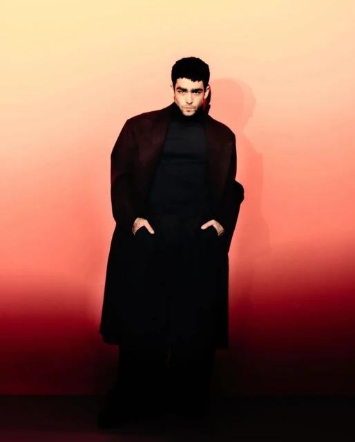 Portrait of a man in a dark turtleneck and long coat, with hands in pockets, posed against a gradient coral and red backdrop.