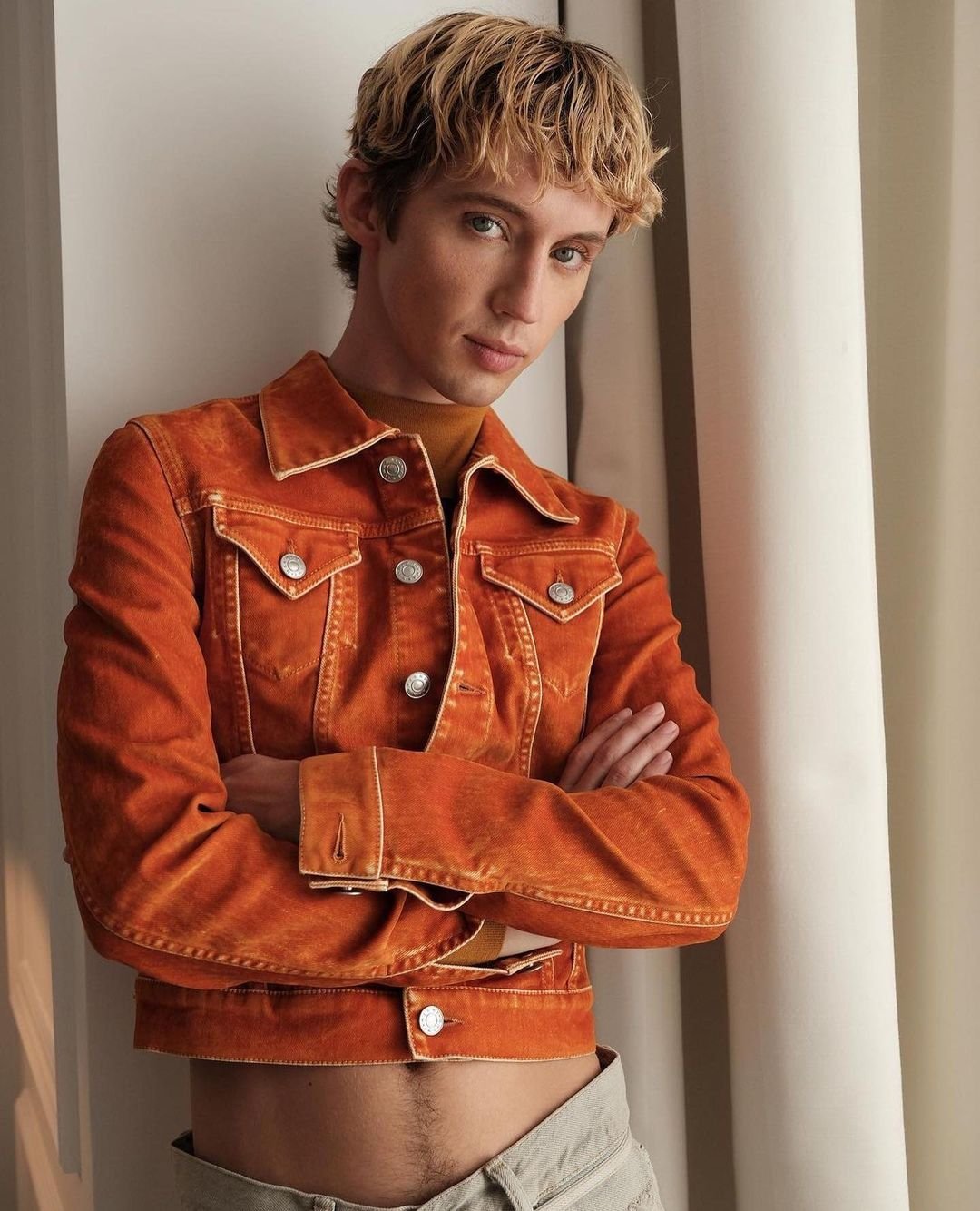 Portrait of a man with blonde curly hair wearing an orange jacket and light grey trousers, leaning against a wall.