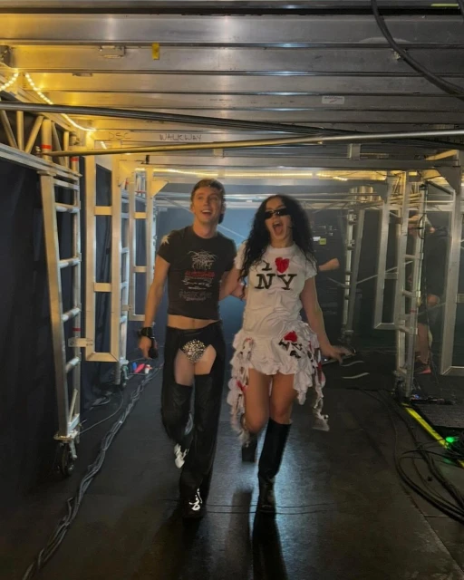 Two people walk through a backstage hallway, one in chaps and a t-shirt, the other in a novelty I Love NY t-shirt dress and boots.