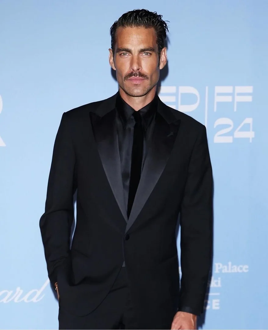 Portrait of an adult male with a mustache in a black suit against a light blue background. Man is wearing a black suit and tie.