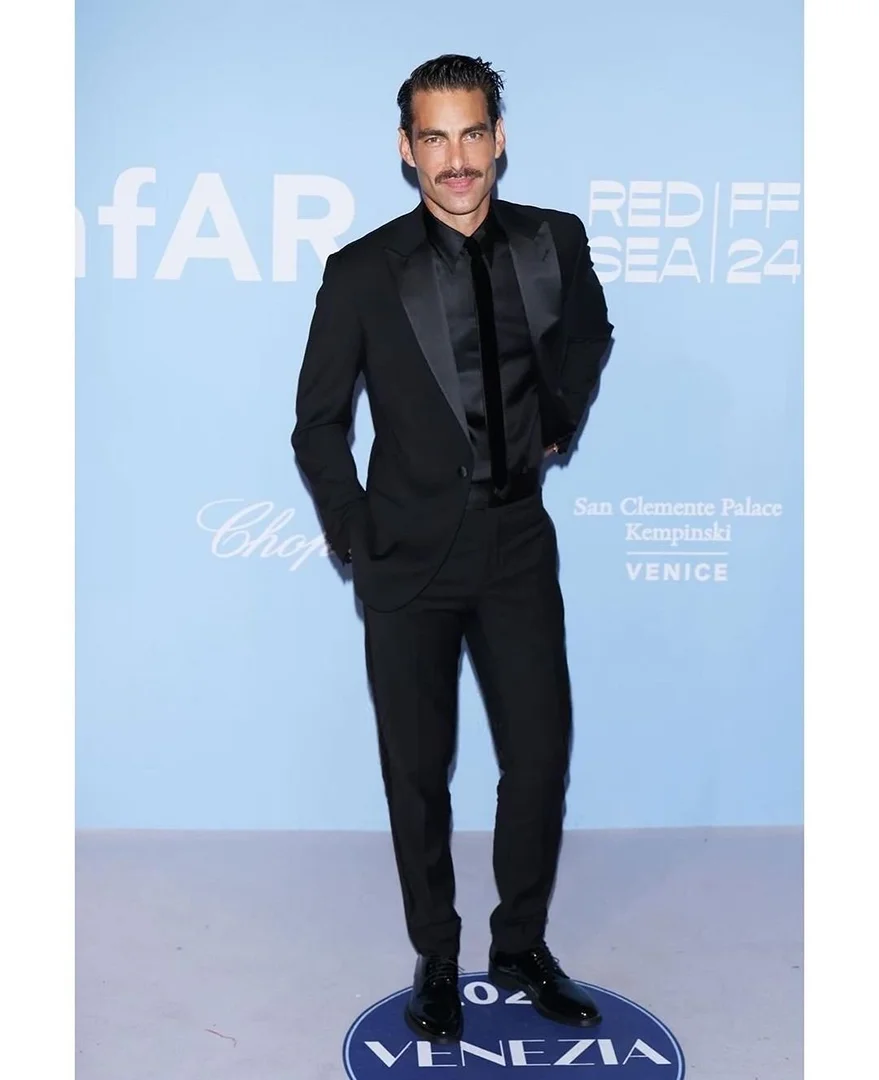 A dapper adult male with a mustache, wearing a black suit and tie, poses at a red-carpet event with a blue and white background.
