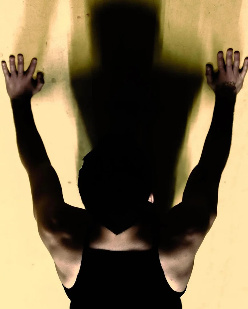 A man in a tank top standing against a tan wall, with his arms and hands raised. A dark shadow of his upper body is cast on the wall.
