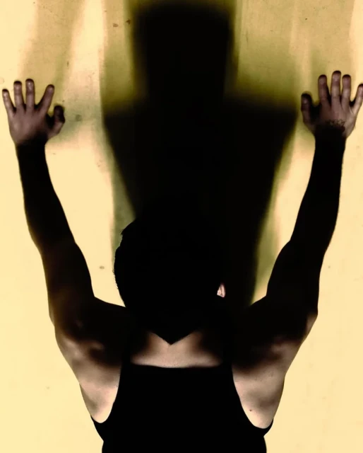 A man in a tank top standing against a tan wall, with his arms and hands raised. A dark shadow of his upper body is cast on the wall.
