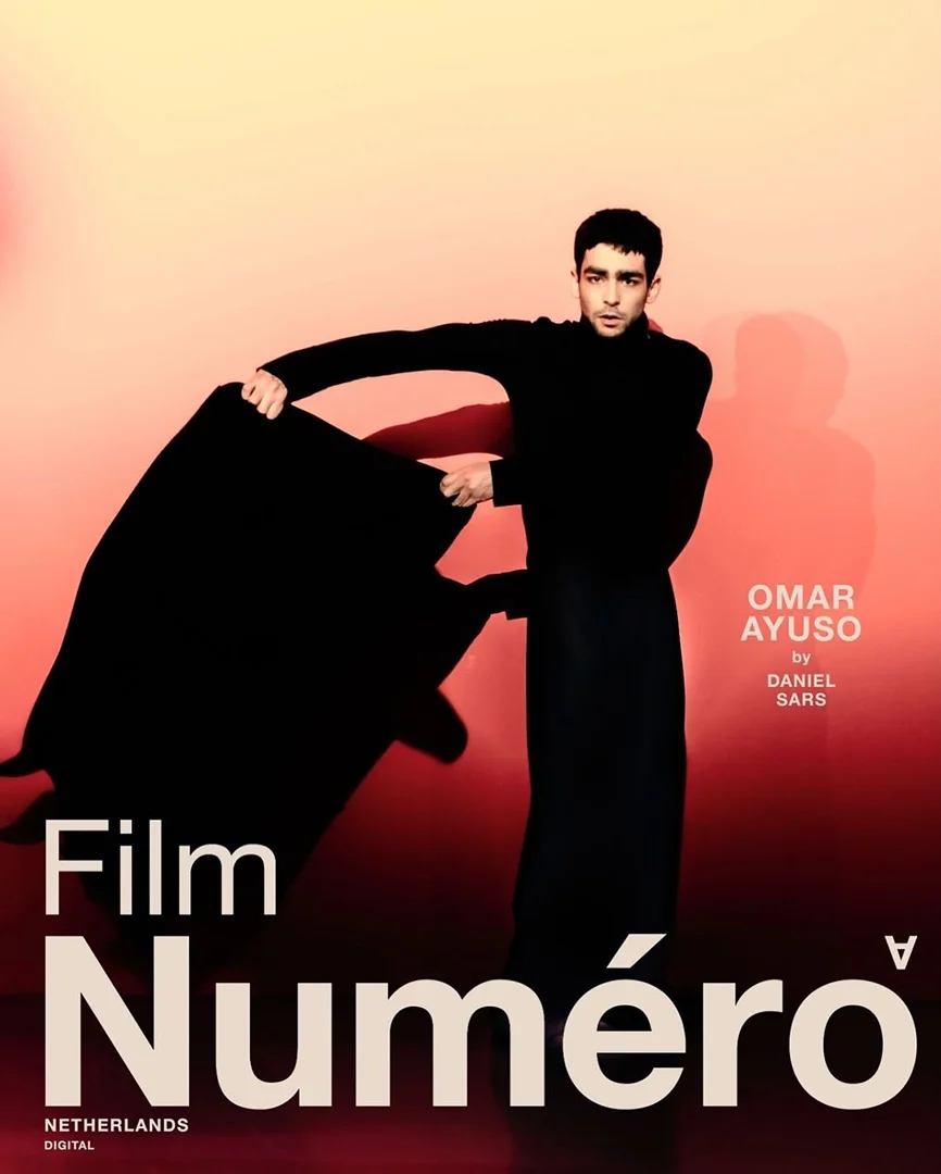 Portrait of a young man in a black outfit, dramatically holding a black fabric against a gradient background in front of magazine cover.