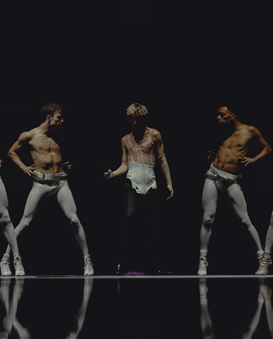 Performance artists on dark stage, dancers with bare torsos, wearing white pants and shoes in a theatrical light setting, dramatic presentation.