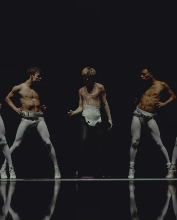 Performance artists on dark stage, dancers with bare torsos, wearing white pants and shoes in a theatrical light setting, dramatic presentation.