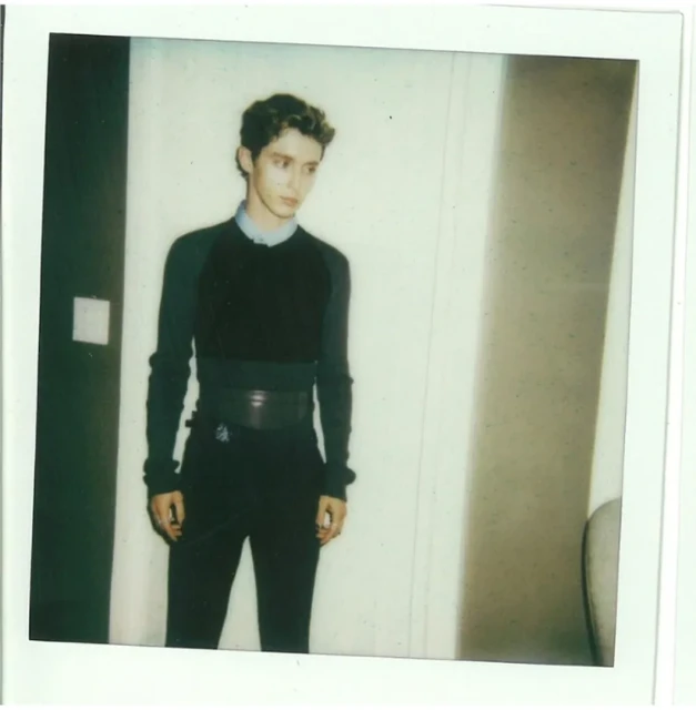 A young man with dark hair poses indoors wearing a black top with long sleeves and black pants with a thick belt, looking off-camera.