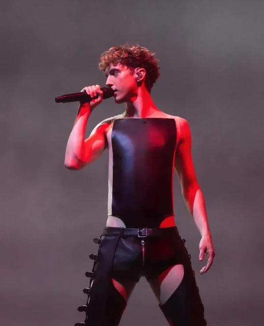 Performer in avant-garde costume singing into a microphone. The individual is standing on stage, spotlit in red, in front of a smoky background.