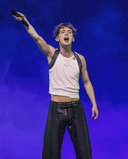 A person in a tank top and leather pants singing into a microphone on a stage with blue lighting at a music concert.