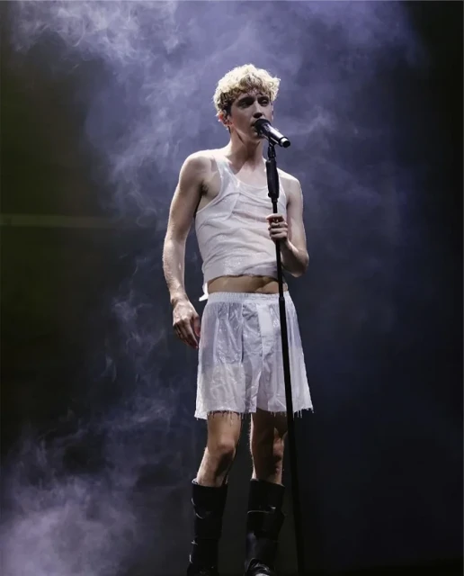 A singer performing on stage with a microphone, wearing a tank top, shorts, and boots, with a smoky background and dark stage lighting.