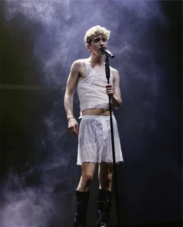 A singer performing on stage with a microphone, wearing a tank top, shorts, and boots, with a smoky background and dark stage lighting.
