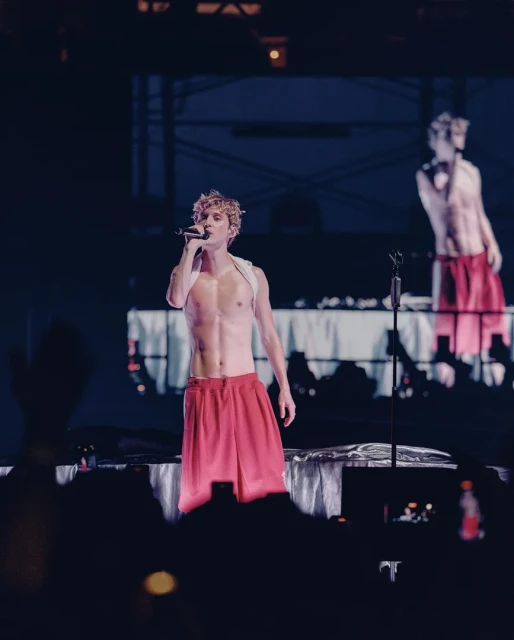 Male singer performing on stage with a microphone, wearing red shorts, shirtless with visible abs, and a white strap across his chest.