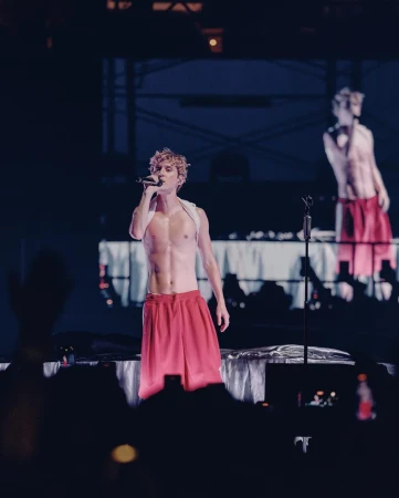 Male singer performing on stage with a microphone, wearing red shorts, shirtless with visible abs, and a white strap across his chest.