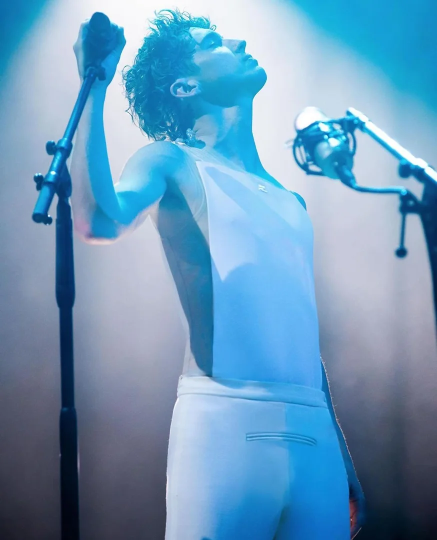 Adult male singer in white outfit performs on stage with microphones and bright blue stage lights, face angled upward in profile shot.