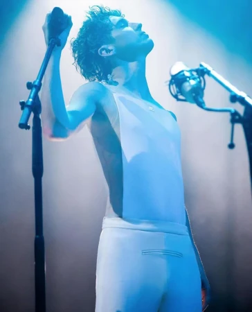 Adult male singer in white outfit performs on stage with microphones and bright blue stage lights, face angled upward in profile shot.