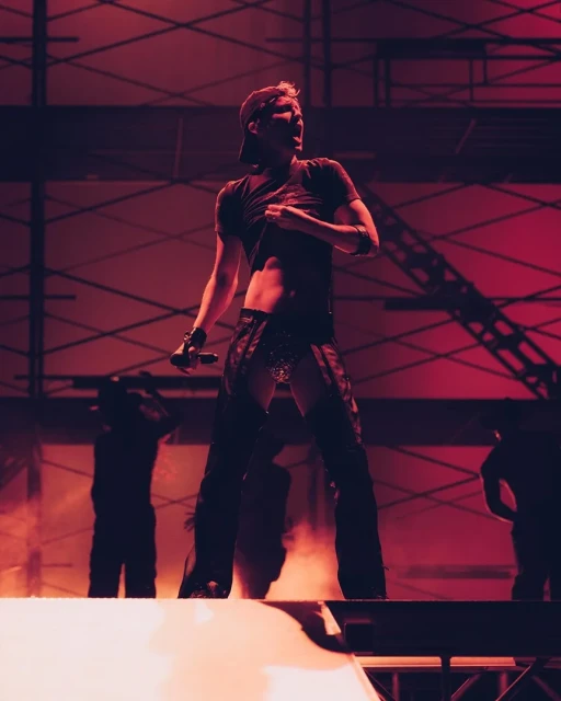 A man is illuminated in red stage lighting, singing and holding a microphone while performing in concert on a stage with metal framework.