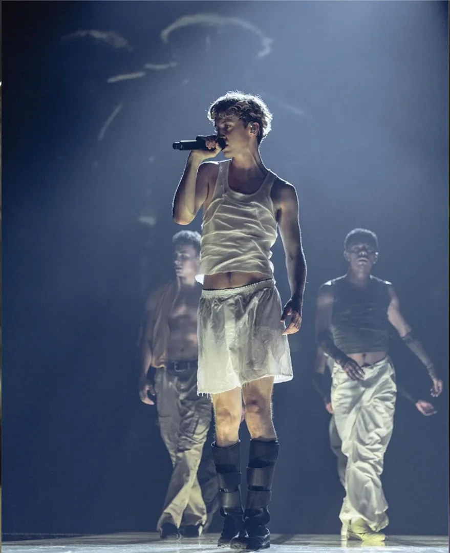 Male performer sings into a microphone on stage in a white tank top with male dancers behind him in a theatrical lighting setup.