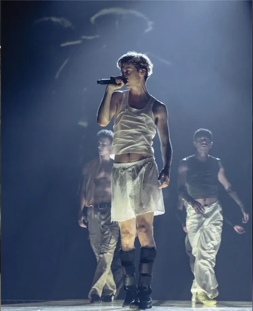 Male performer sings into a microphone on stage in a white tank top with male dancers behind him in a theatrical lighting setup.