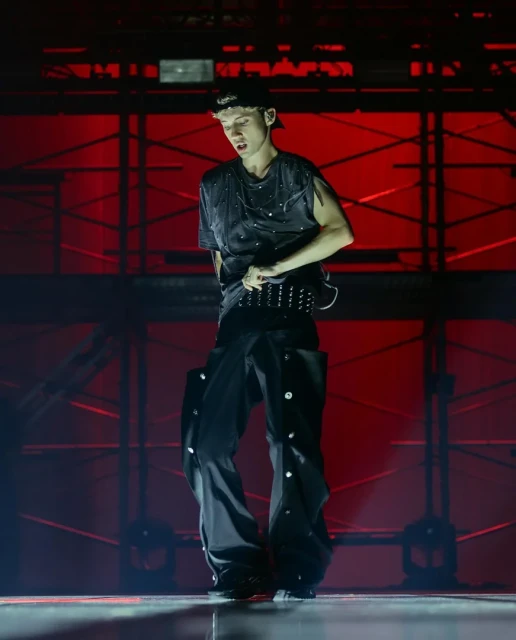 Performer in a black outfit and cap stands on stage with red lighting and geometric background structure during a concert.