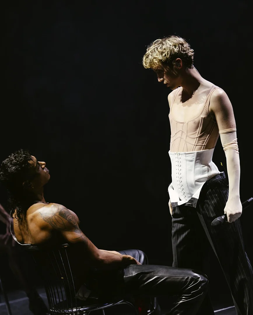 Two people on stage: One seated in a chair and wearing leather pants, the other standing and wearing a corset and pinstripe pants.