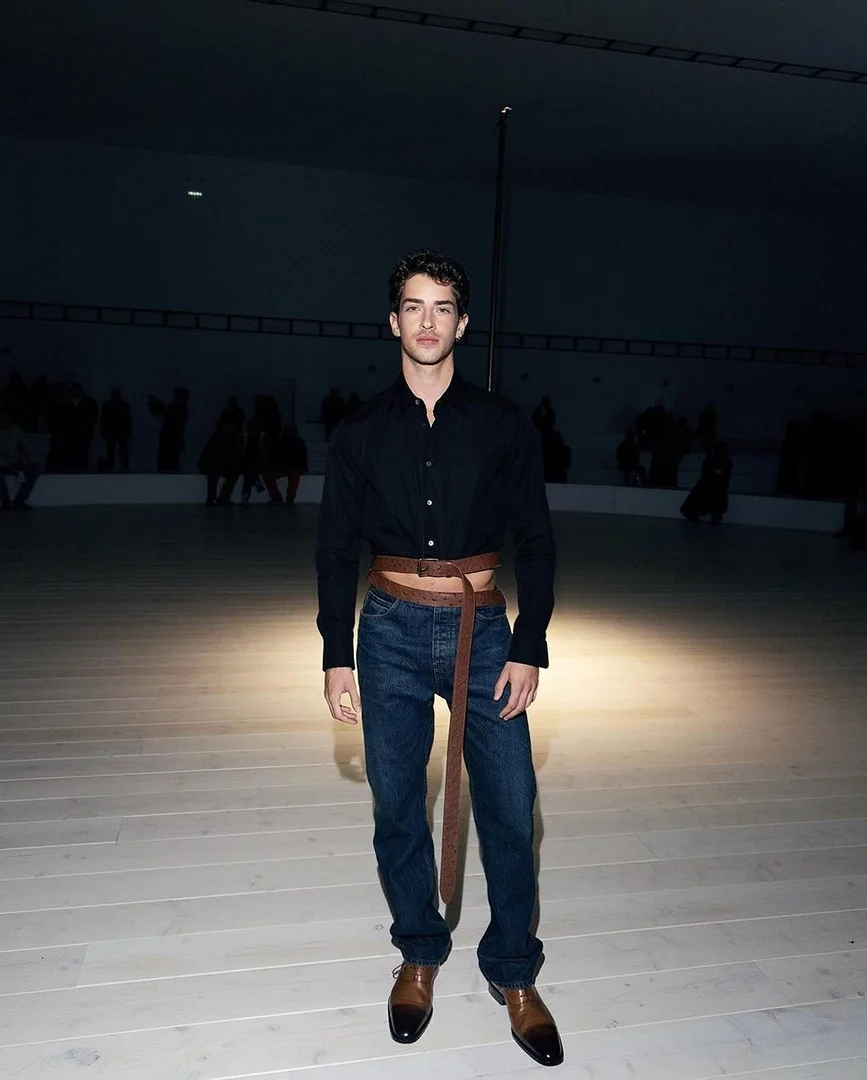 Man in button-down shirt and jeans poses in a spacious room with a light floor and dim lighting, creating a fashion-forward and stylish look.