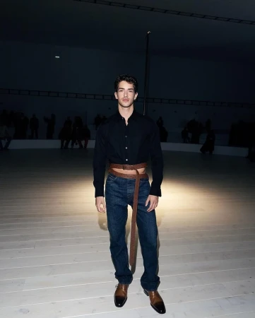 Man in button-down shirt and jeans poses in a spacious room with a light floor and dim lighting, creating a fashion-forward and stylish look.