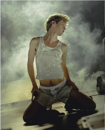 Young man kneeling on stage wearing a white tank top and shorts amidst theatrical stage smoke styled by Marc Forne