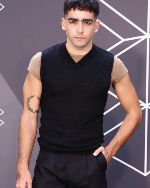 Portrait of an adult male with dark hair and a black vest posing against a gray background with geometric white lines.