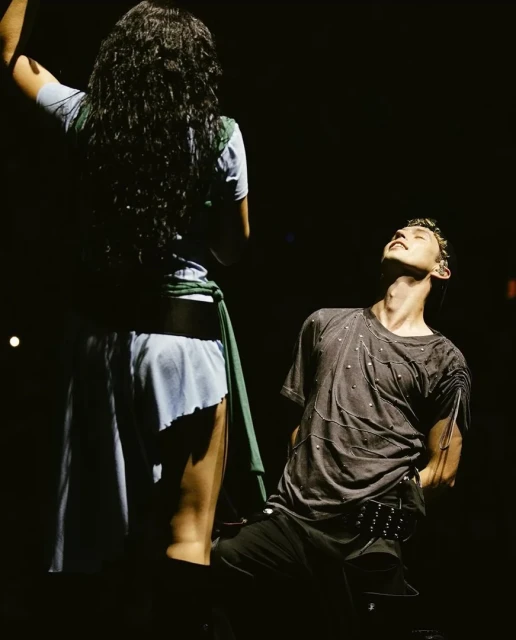 Two figures on a dark stage, illuminated by spotlights. A person with long, dark hair is standing next to an individual with head tilted back.