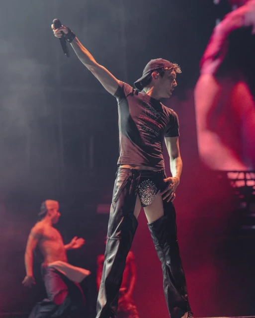 A male entertainer with a microphone sings on stage during a live performance in black leather chaps with dancers in the background.