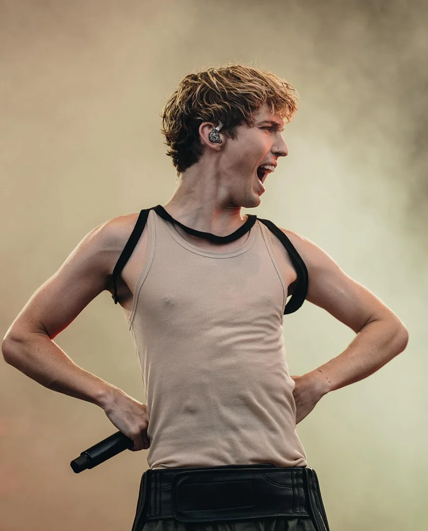 Singer performs on stage with microphone, wearing a light tank top and black harness; arms are raised with an open mouth expression.
