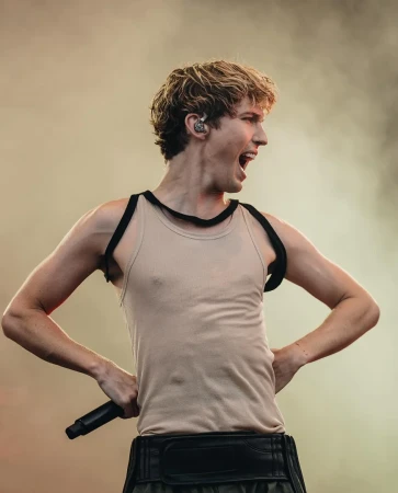 Singer performs on stage with microphone, wearing a light tank top and black harness; arms are raised with an open mouth expression.