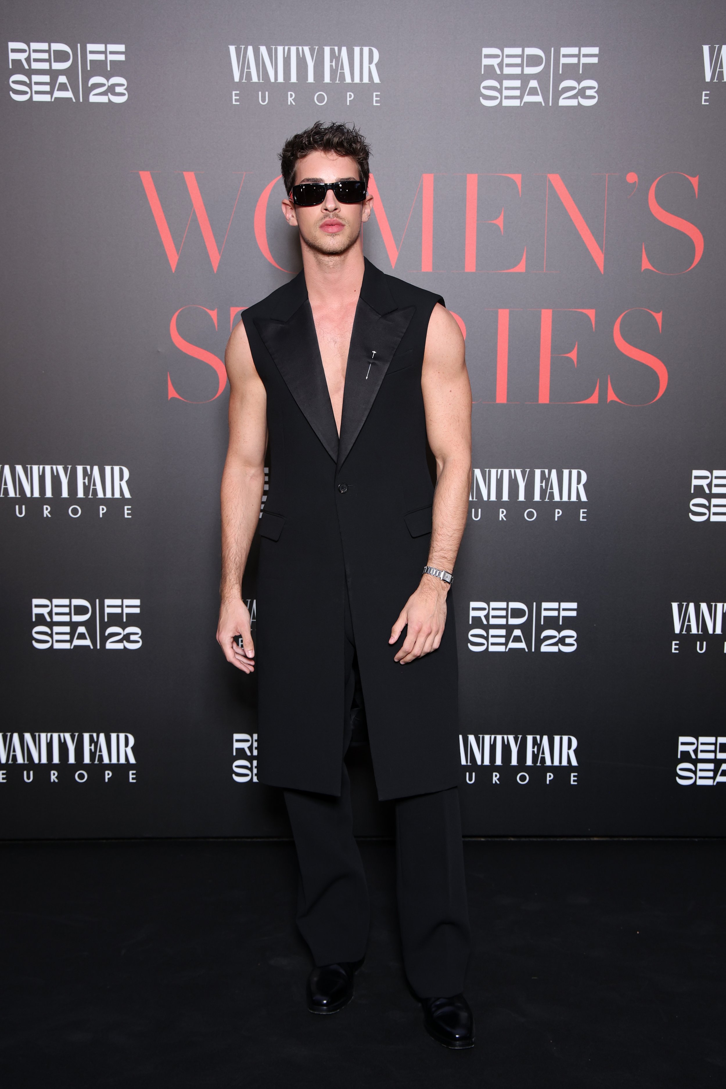 Man posing in long sleeveless tuxedo jacket at REDIFF SEA 23 and Vanity Fair Europe event with Women's Series text in background.