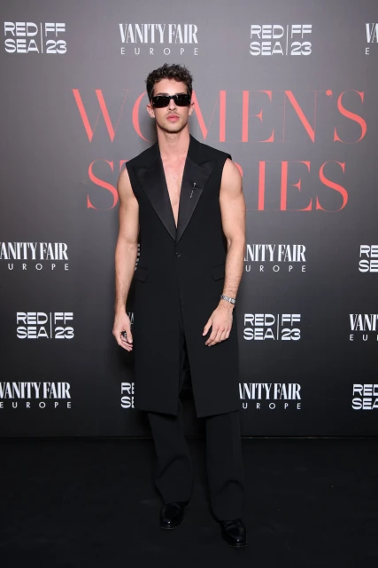 Man posing in long sleeveless tuxedo jacket at REDIFF SEA 23 and Vanity Fair Europe event with Women's Series text in background.