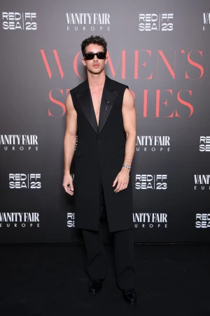 Man posing in long sleeveless tuxedo jacket at REDIFF SEA 23 and Vanity Fair Europe event with Women's Series text in background.
