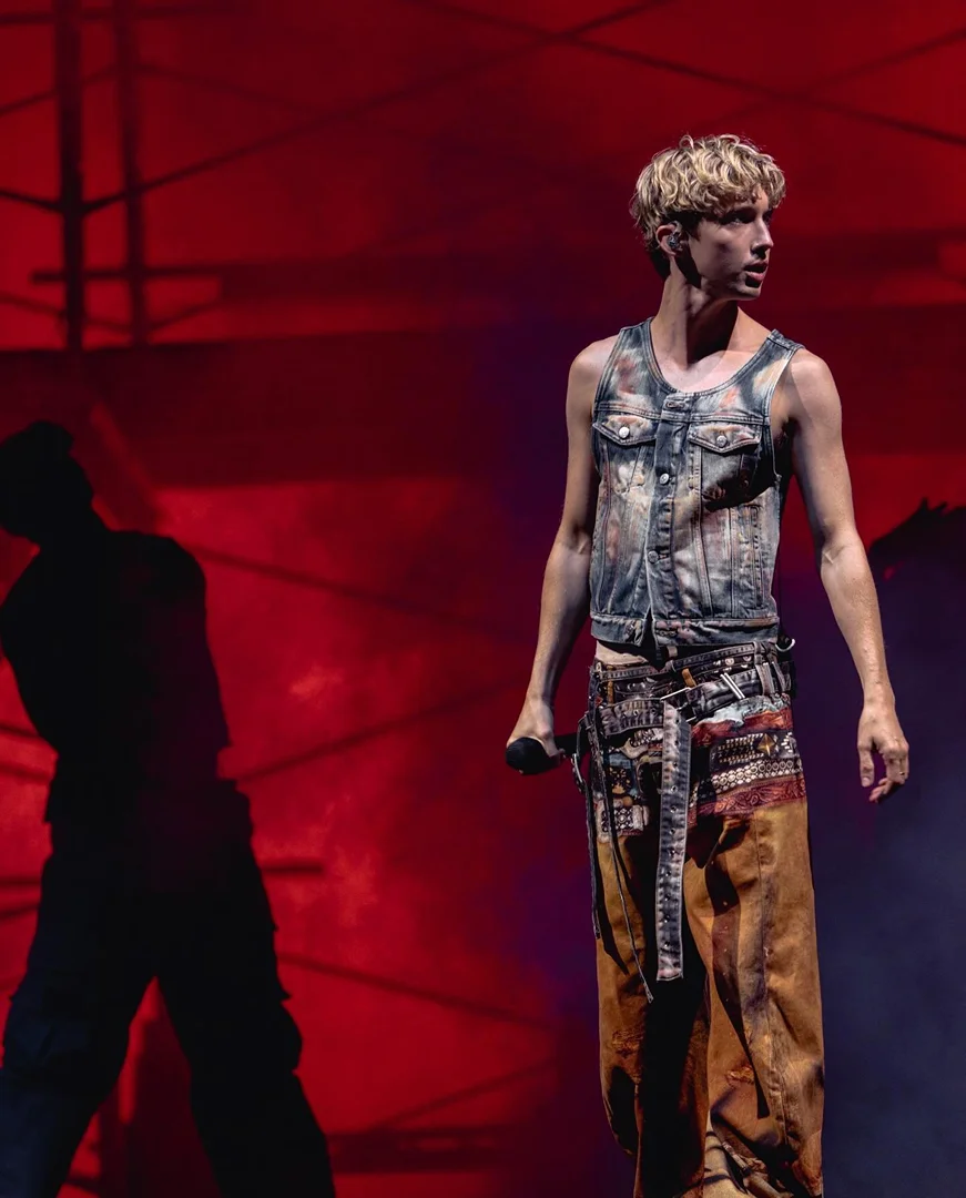A young adult male musician stands on a stage with a microphone, wearing a denim vest and patterned pants, with a red backdrop.