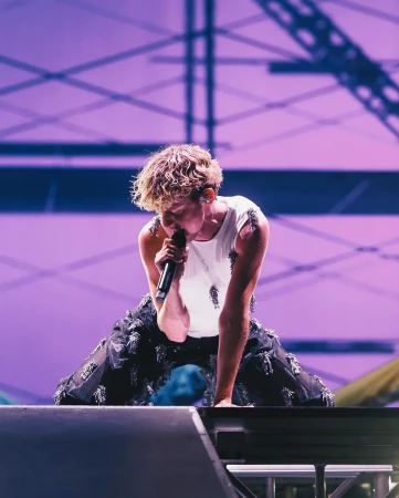 A singer performing on stage, kneeling while holding a microphone, with a purple-lit background and visible stage equipment.
