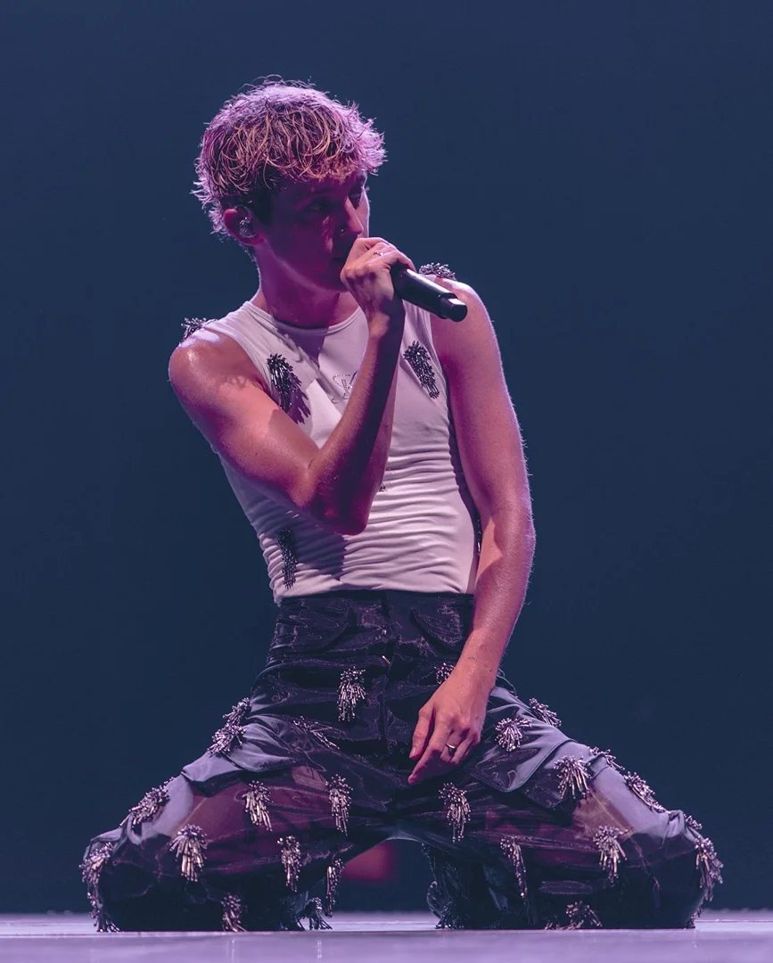 A male singer kneels on stage, holding a microphone. He is wearing a white tank top and dark, sheer trousers embellished with silver details.