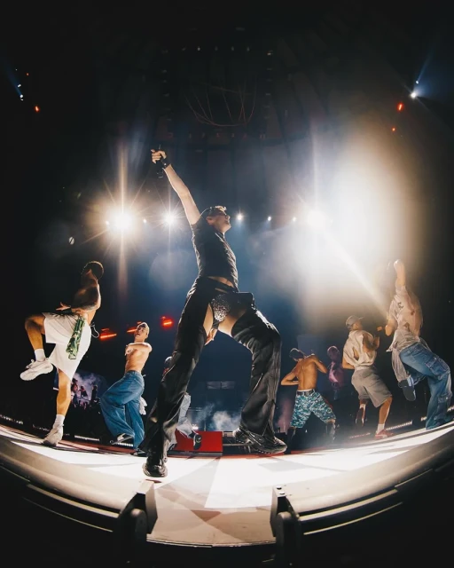 A male singer with dancers performs on a concert stage with bright lights, wide angle lens, and a concert venue background.