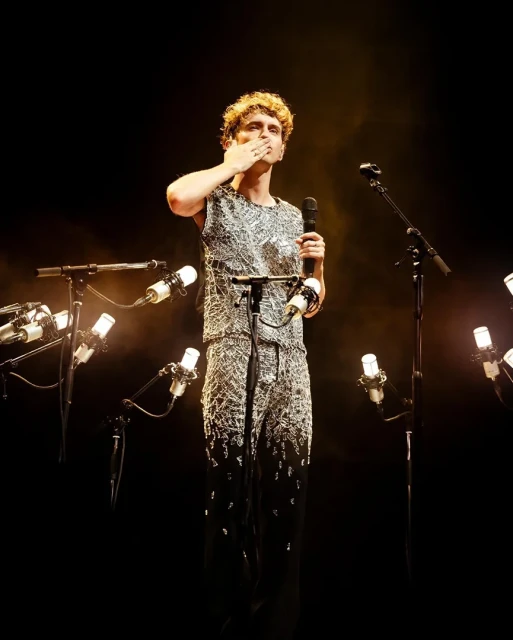 Male singer performing on stage in a silver outfit, blowing a kiss to the audience with microphones and stage lighting in the background.