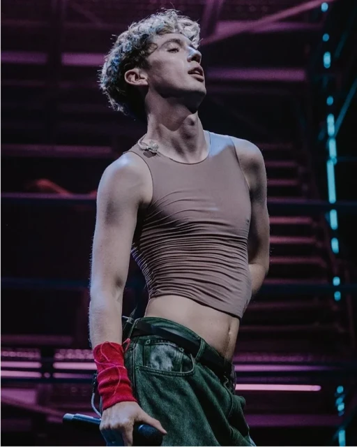 Adult male singer performs on stage wearing a brown tank top and baggy jeans under vibrant neon lighting, holding a microphone.